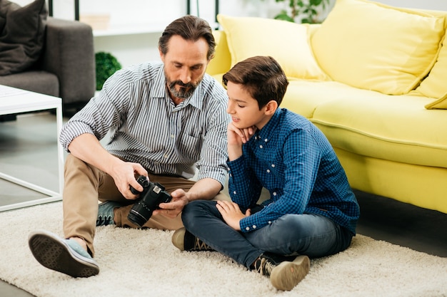 Neugieriger dunkelhaariger Junge, der lächelt, während er mit seinem Vater auf dem Teppich sitzt und Fotos auf dem Bildschirm seiner Kamera betrachtet