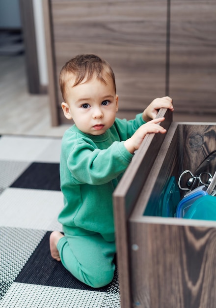 Neugieriger kleiner Junge, der die Schublade in der Küche erkundet Das kleine Baby sitzt auf dem Küchenboden Kindersicherheitskonzept