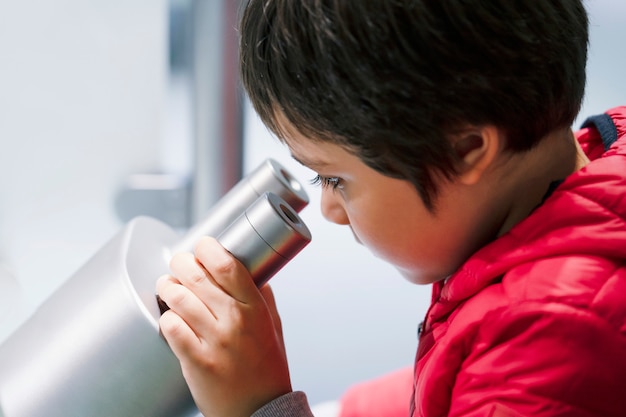 Neugieriger kleiner Junge, der durch Mikroskop beim Haben des Spaßes im wissenschaftlichen Verein nach Vorschülern sucht