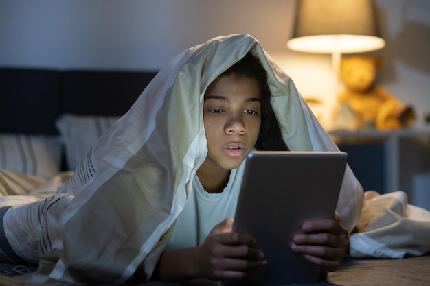 Neugieriges afroamerikanisches Mädchen, das unter der Bettdecke im Bett liegt und nachts auf dem Tablet surft?