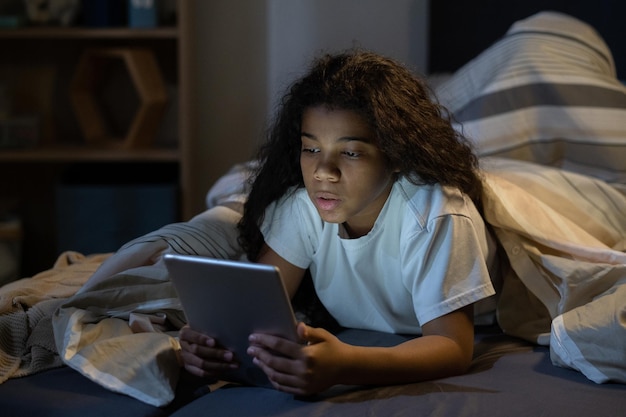 Neugieriges afroamerikanisches Mädchen mit welligem Haar, das unter der Bettdecke im Bett liegt und Tablet im dunklen Raum benutzt