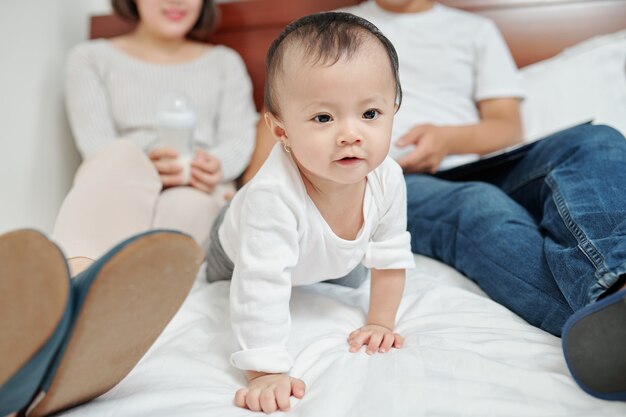 Neugieriges kleines asiatisches Mädchen, das kriecht, wenn ihre Eltern nach langem Tag auf Bett ruhen