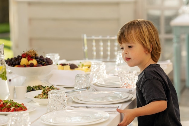 Foto neugieriges kleinkind, das einen festlichen tisch beobachtet, der exquisit für ein bankett auf einer weißen tischdecke serviert wird und auf gäste wartet