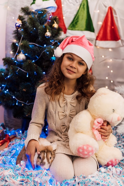 Neujahr ein Mädchen in einer rosa Mütze hält ein Kaninchen und einen Teddybären in der Nähe des Weihnachtsbaumes