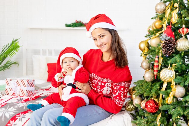 Neujahr oder Weihnachten, eine junge Mutter mit einem Baby auf dem Bett zu Hause am Weihnachtsbaum in einem Weihnachtsmann-Kostüm umarmt und wartet auf den Urlaub lächelnd