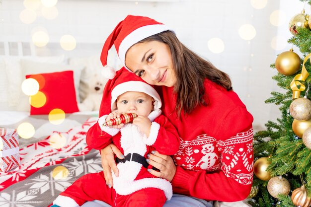 Neujahr oder Weihnachten, eine junge Mutter mit einem Baby auf dem Bett zu Hause am Weihnachtsbaum in einem Weihnachtsmann-Kostüm umarmt und wartet auf den Urlaub lächelnd