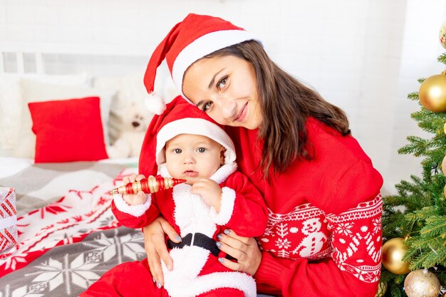 Neujahr oder Weihnachten, eine junge Mutter mit einem Baby auf dem Bett zu Hause am Weihnachtsbaum in einem Weihnachtsmann-Kostüm umarmt und wartet auf den Urlaub lächelnd