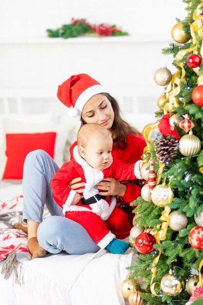 Neujahr oder Weihnachten, eine junge Mutter mit einem Baby auf dem Bett zu Hause am Weihnachtsbaum in einem Weihnachtsmann-Kostüm umarmt und wartet auf den Urlaub lächelnd