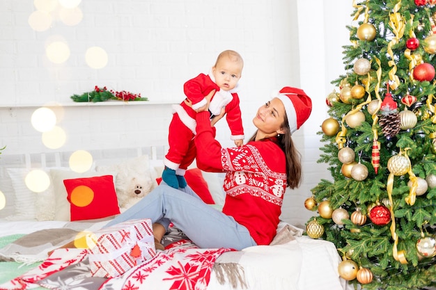 Neujahr oder Weihnachten, eine junge Mutter mit einem Baby auf dem Bett zu Hause am Weihnachtsbaum in einem Weihnachtsmann-Kostüm umarmt und wartet auf den Urlaub lächelnd