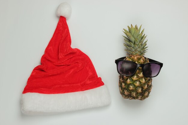 Neujahr, Weihnachtskonzept. Ananas gekleidet in Gläsern mit Weihnachtsmütze auf weißem Hintergrund. Draufsicht.