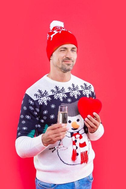 Neujahrs-Herzgeschenk und Mann im Winterpullover Mann mit Neujahrshut mit Champagner und Herz Neujahrsmann isoliert auf rotem Hintergrund Mann hält Champagner für Neujahr im Studio