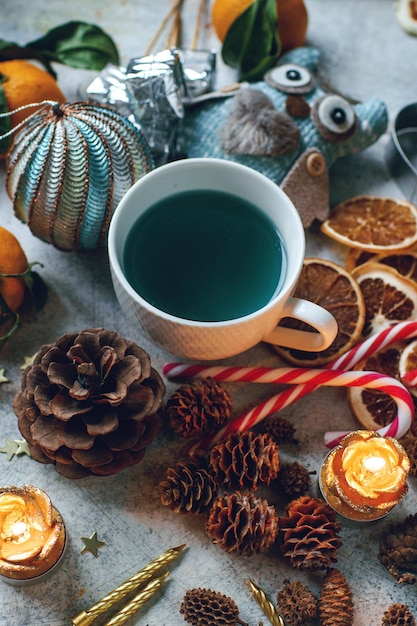 Neujahrs- oder Weihnachtskomposition mit blauem Tee, Mandarinen, trockenen Orangenkegeln, Zimtgeschenken, Kerzen, Süßigkeiten und Spielzeug auf hellem rustikalem Tisch