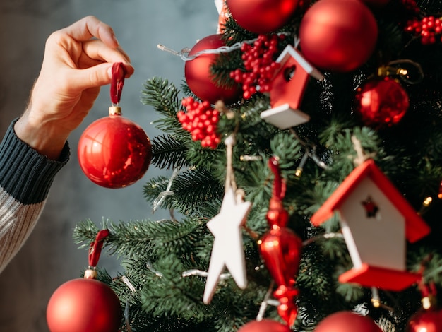 Neujahrsfeierkonzept. Nahaufnahme der Mannhand, die rote Kugelverzierung hält, um grünen Tannenbaum zu verzieren.