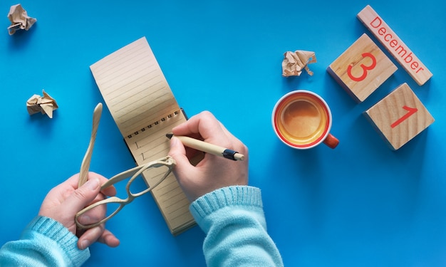 Neujahrsvorsatzkonzept, Hand mit Kaffee, Notizbuch, Holzkalender und Kaffee auf Blau