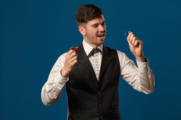 Neuling im Poker in schwarzer Weste und weißem Hemd mit farbigen Chips, die vor blauem Hintergrund posieren