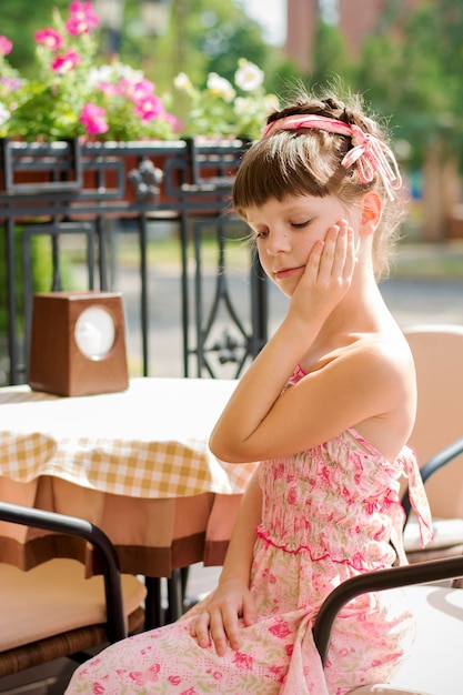 Neunjähriges Mädchen mit geflochtenen Haaren sitzt an einem Tisch in einem Café auf der Sommerterrasse. Traurige Blicke legen ihre Hand auf ihre Wange