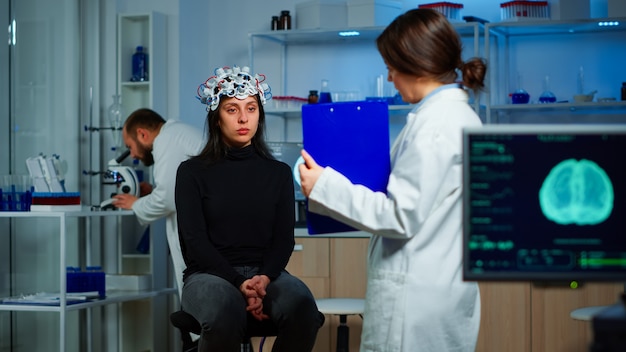 Neurowissenschaftlicher Arzt mit Zwischenablage, der Patienten mit EEG-Headset Behandlung gegen Hirnerkrankungen zeigt. Frau, die im neurologischen wissenschaftlichen Labor sitzt und Funktionsstörungen des Nervensystems behandelt