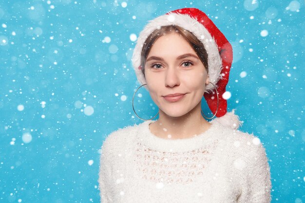 Neve, inverno, natal, pessoas, conceito de beleza - loira e bonita usando chapéu de natal em fundo de neve azul claro. retrato de uma jovem linda e alegre menina sorrindo, olhando para a câmera