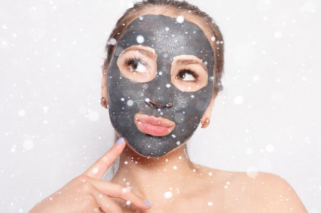 Neve, inverno, Natal, pessoas, conceito de beleza - máscara facial de mulher. Retrato de uma linda garota removendo a máscara cosmética preta da pele do rosto sobre um fundo de neve