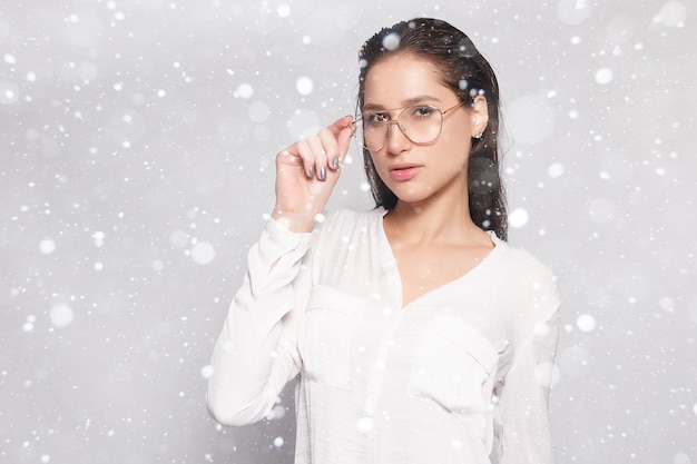 Neve, inverno, Natal, pessoas, conceito de beleza - muito jovem em t-shirt branca em branco, usando óculos, parede vazia, retrato de estúdio. Mulher morena sorridente com óculos sobre fundo de neve