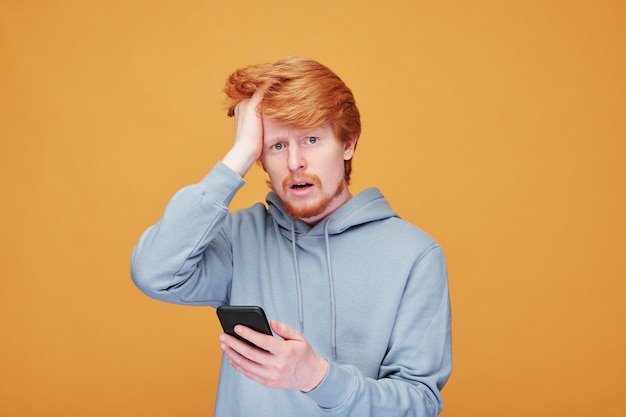 Ängstlicher junger Mann mit roten Haaren, die den Kopf berühren und die Vorderseite gegen die gelbe Wand betrachten, während sie aktuelle Nachrichten im Mobiltelefon lesen