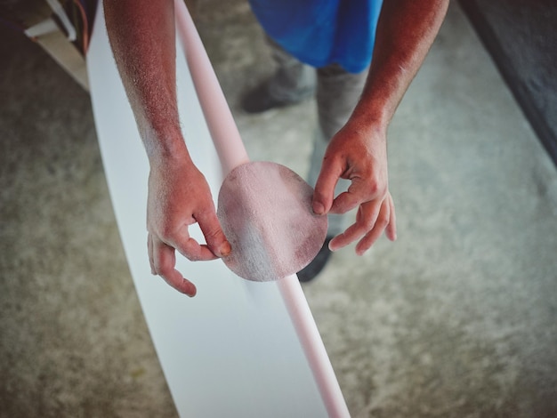 Ángulo alto de cultivo tatuado trabajador masculino anónimo lijando tabla de surf mientras trabaja en el taller de artesanía