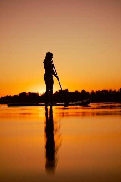 Ângulo baixo da senhora de meia idade embarcando no lago com o sol se pondo abaixo do horizonte logo atrás de seu corpo com reflexo na superfície da água Estilo de vida ativo ao ar livre