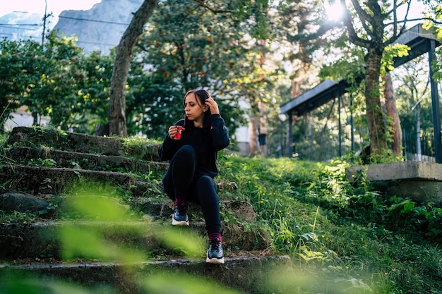 Ángulo bajo de joven viajera en ropa activa sentada en viejas escaleras de piedra y tomando café mientras descansa durante la caminata en el parque botánico verde