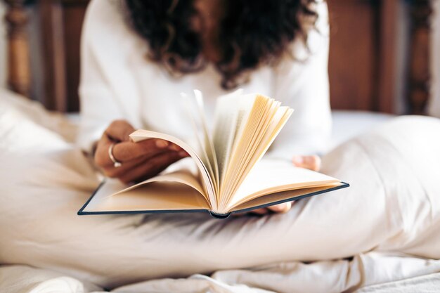 Nicht erkennbare junge Frau, die die Seiten eines Buches umblättert, während sie im Bett liest, das mit der Bettdecke bedeckt ist