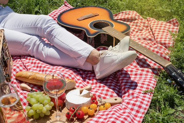 Nicht erkennbare junge Frau in weißen Hosen draußen, die Picknick isst und Gitarre spielt