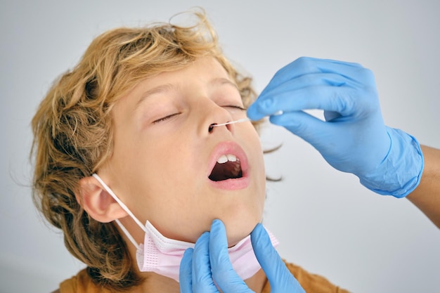 Nicht erkennbare Krankenschwester mit Tupfer, der den COVID-19-Test aus der Nase eines Kindes mit geschlossenen Augen auf hellem Hintergrund durchführt