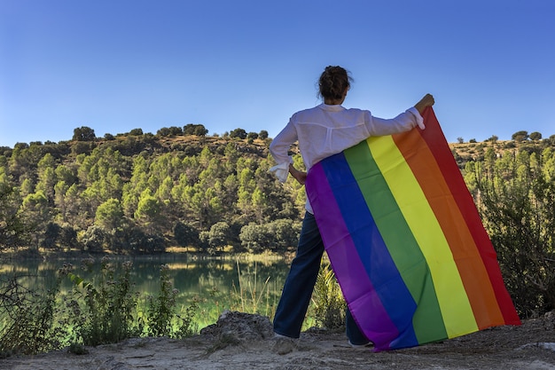 Nicht erkennbare lesbische Frau mittleren Alters, die die Gay Rainbow Flag am See im Freien hält. Freiheitskonzept