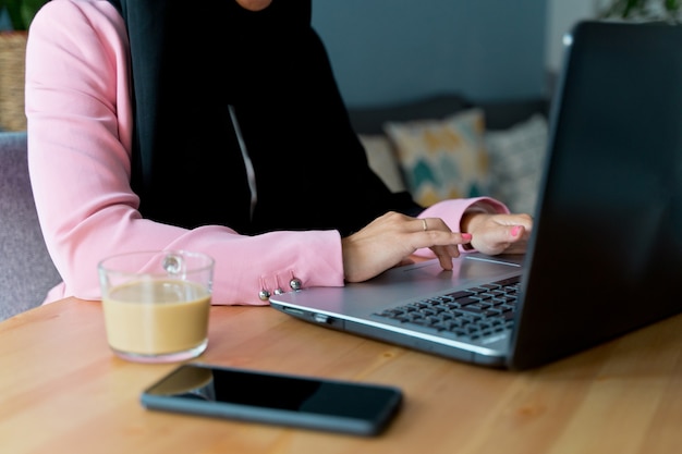 Nicht erkennbare muslimische Frau, die einen Hijab mit Technologie im Innenbereich trägt. Horizontale Seitenansicht der arabischen Frau, die zu Hause mit Laptop arbeitet. Technologie und Lebensstil der muslimischen Frauen.