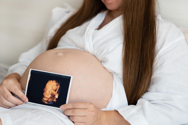 Nicht erkennbare schwangere Frau, die zu Hause Robe trägt, streichelt, streichelt den Bauch, Babybauch, hält Ultraschall-Sonogramm-Foto, abgeschnittene Nahaufnahme, glücklich, natürlich, ruhig, gesunde Schwangerschaft