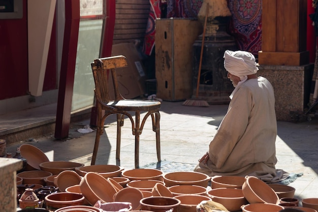 Nicht erkennbarer alter arabischer Mann mit traditioneller Kleidung, der handgefertigte Tonschalen auf dem Straßenmarkt verkauft