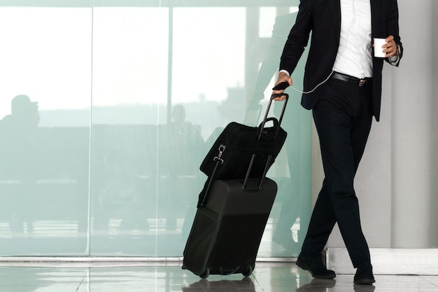 Nicht erkennbarer Geschäftsmann, der mit seinem Koffer durch den Flughafen geht, während er einen Kaffee in der Hand hält