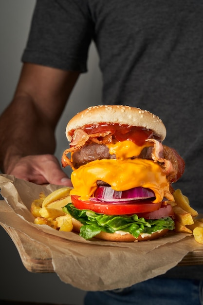 Nicht erkennbarer junger Mann, der einen Holztisch mit leckeren schnellen Burgern und Pommes auf grauem Hintergrund hält