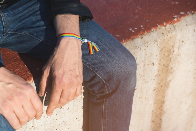 Nicht erkennbarer Mann in Jeans und schwarzer Lederjacke, die auf einer Betonbank mit Regenbogen-Schwulenstolzarmband sitzen