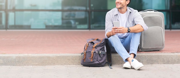 Nicht erkennbarer Mann sitzt mit Gepäck in der Nähe des Flughafenterminals und benutzt Smartphone