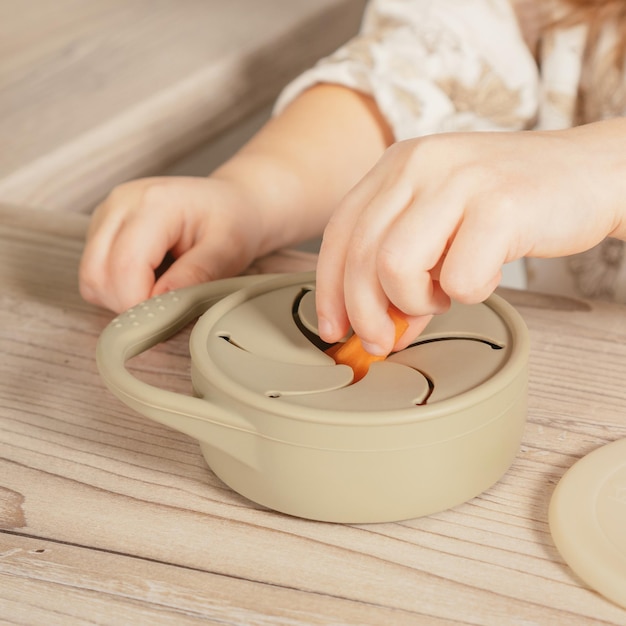 Nicht erkennbares Kind, das ein Stück Karotte aus dem pastellgrauen Silikon-Snackbecher in der Nähe des Deckels am Holztisch nimmt Babyzubehör Geschirr erstes Fütterungskonzept Draufsicht Instagram verwendet quadratischen Rahmen