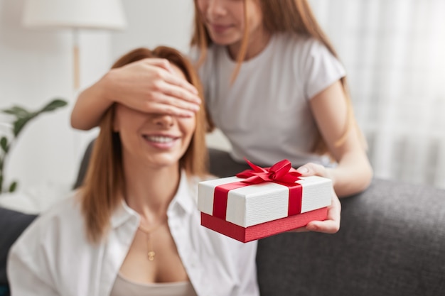 Nicht erkennbares Teenager-Mädchen mit Geschenkbox, die die Augen der lächelnden Mutter bedeckt, während sie Überraschung macht und zum Geburtstag zu Hause gratuliert