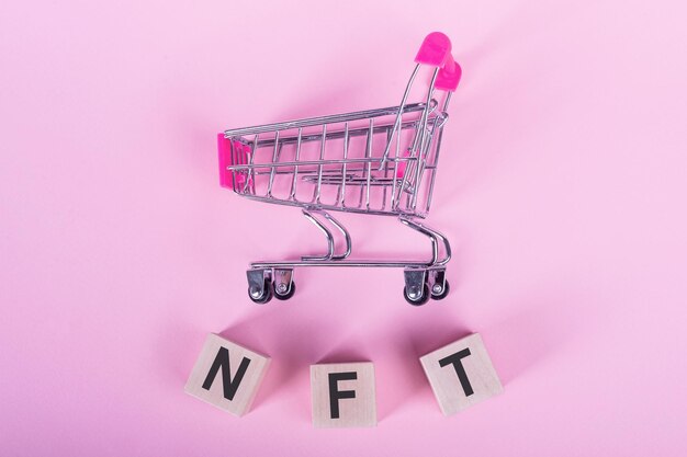 Nicht fungibles NFT-Token-Wort auf Holzwürfeln auf rosafarbenem Hintergrund mit einem Einkaufswagen