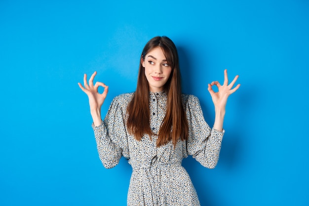 Nicht schlecht. Erfreut lächelnde Frau mit langen Haaren, Kleid, zeigt gute Zeichen und sieht zufrieden aus, lobt gute Arbeit, schaut links auf Logo, blau.