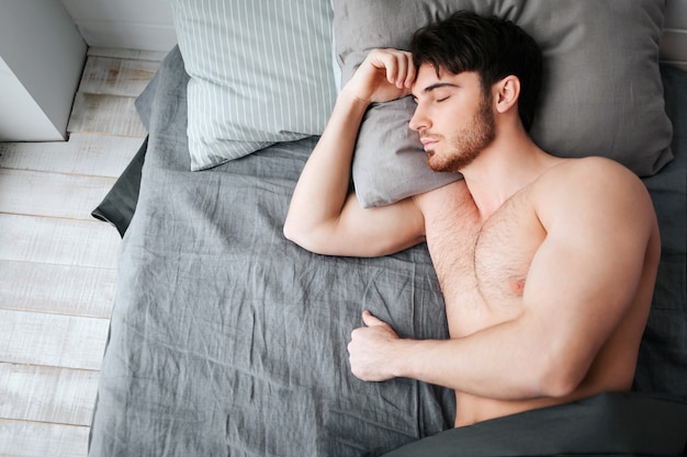 Único homem musculoso jovem dormindo na cama. Ele segura a cabeça no travesseiro. Jovem está nu. Parte inferior do corpo coberta com manta cinza.