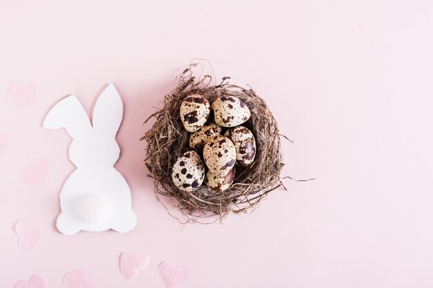 Nido de pascua feliz con huevos de codorniz y conejo de cartón blanco sobre fondo rosa vista superior