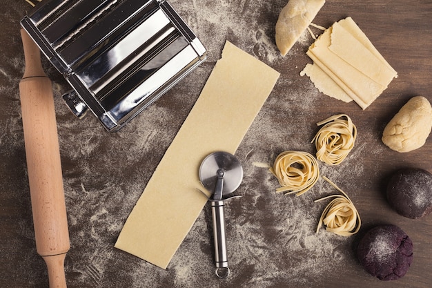 Nido de tallarines crudos y rebanadas de masa con cortador de rueda en la mesa de la cocina. Vista superior de ingredientes y equipos para preparar pasta italiana, espacio de copia