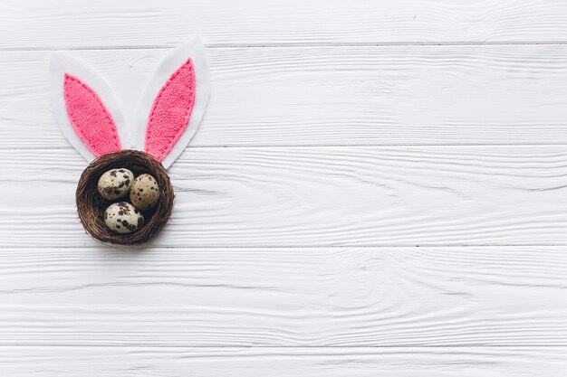 Nido con tres huevos de Pascua rosa y orejas de conejo en el fondo blanco de madera.