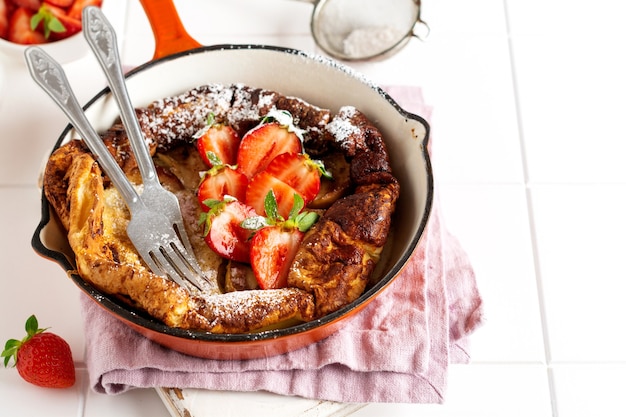 Niederländischer Babypfannkuchen mit frischer Erdbeerbeere und mit Puderzuckerpulver in roter Pfanne auf weißer Küchenoberfläche bestreut. Draufsicht.