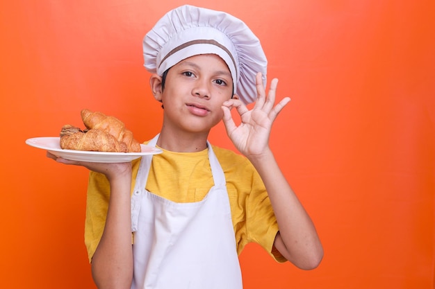 Niedlicher, entzückender kleiner Junge in Kochuniform hält einen Teller mit Croissantbrot und zeigt ein köstliches Schild