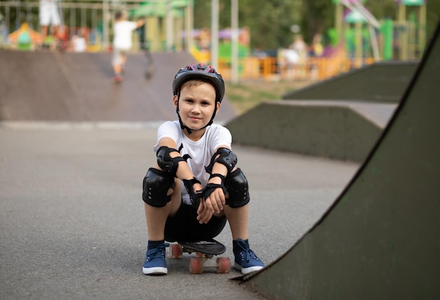 Niedlicher kleiner Junge mit Helm sitzt in einem speziellen Bereich im Skatepark und sitzt auf dem Skateboard Sommersport-Aktivitätskonzept Glückliche Kindheit